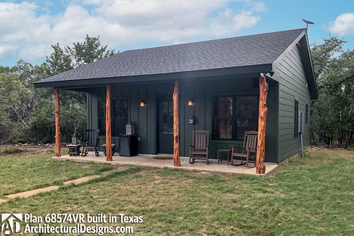 House plan exterior with a deep covered porch supported by rustic log posts, dark green siding, and gabled roof.