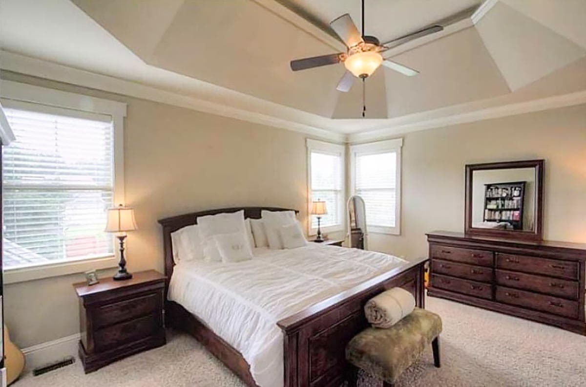 Interior view of a bedroom with a tray ceiling, ceiling fan, dark wood furniture, and multiple windows.