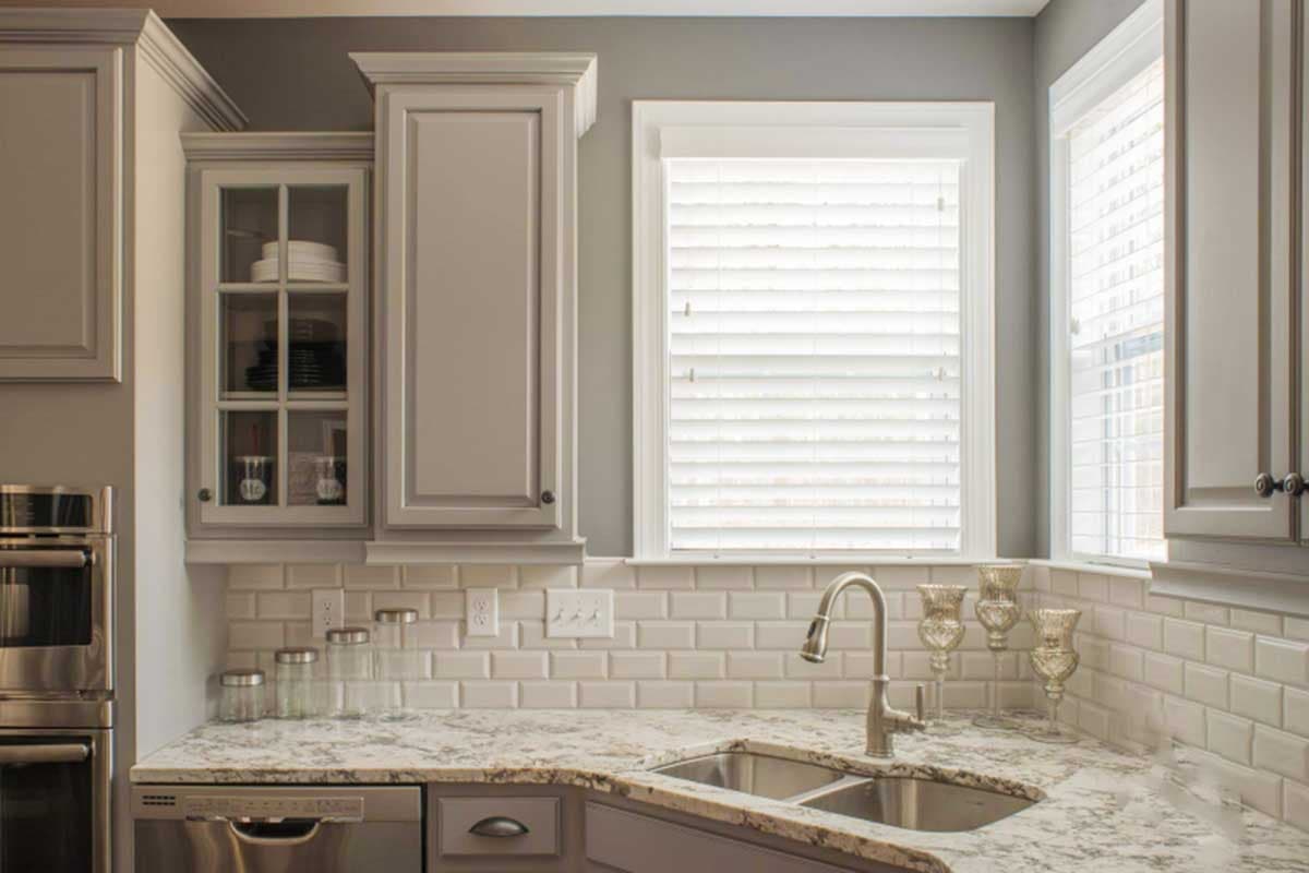 Kitchen interior with gray cabinets, subway tile backsplash, granite countertops, stainless steel sink, and double windows with blinds.