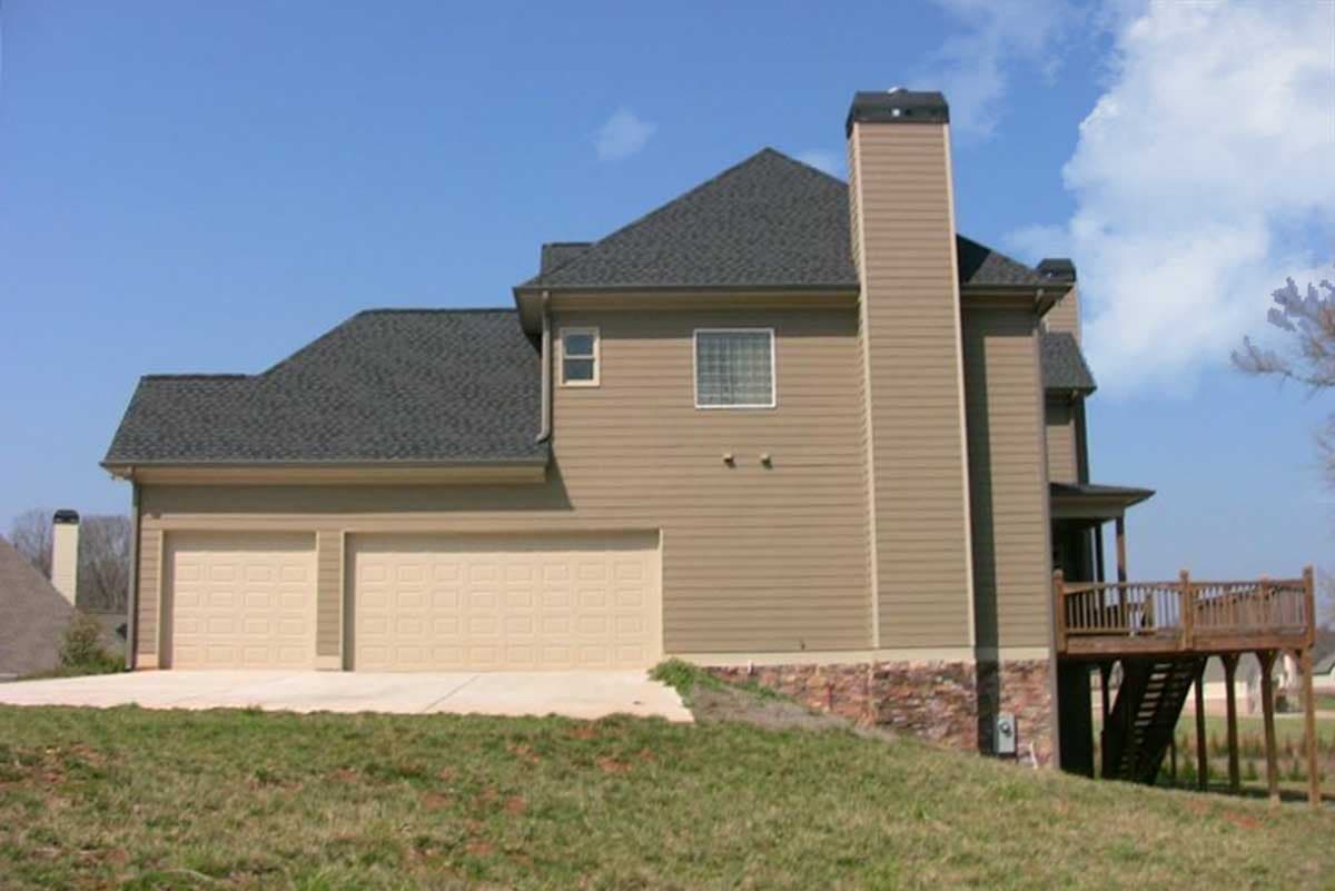 House plan exterior: two-story home with a side-facing three-car garage, large stone chimney, and elevated wooden deck.