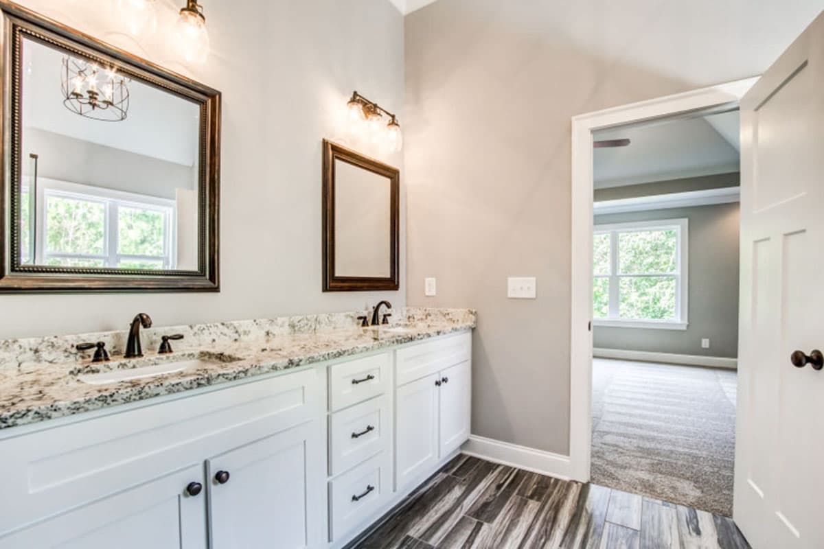 Bathroom vanity with double sinks, granite countertops, and wood-look tile flooring. View into bedroom.