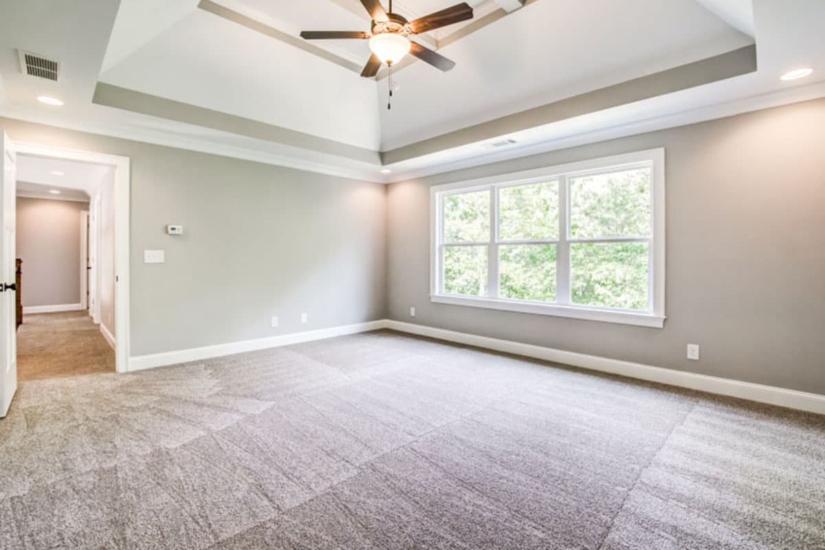 Interior view of a bedroom with tray ceiling, ceiling fan, large multi-pane window, and carpeted floor. Hallway visible.