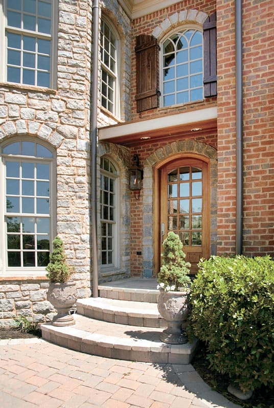 House plan exterior: Stone and brick facade with arched windows, wooden shutters, and a multi-pane glass entry door.