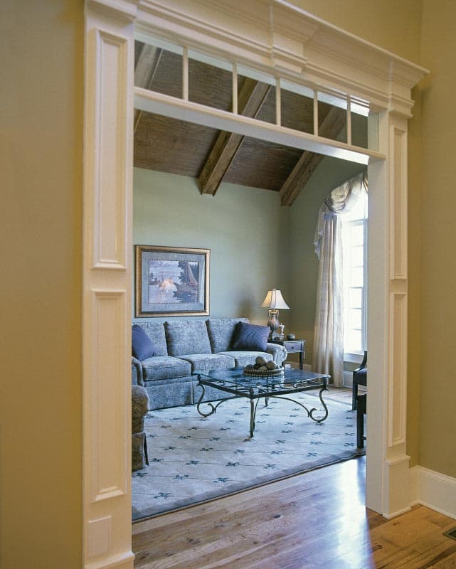 Living room with vaulted ceiling, exposed beams, patterned sofa, glass coffee table, and large window.