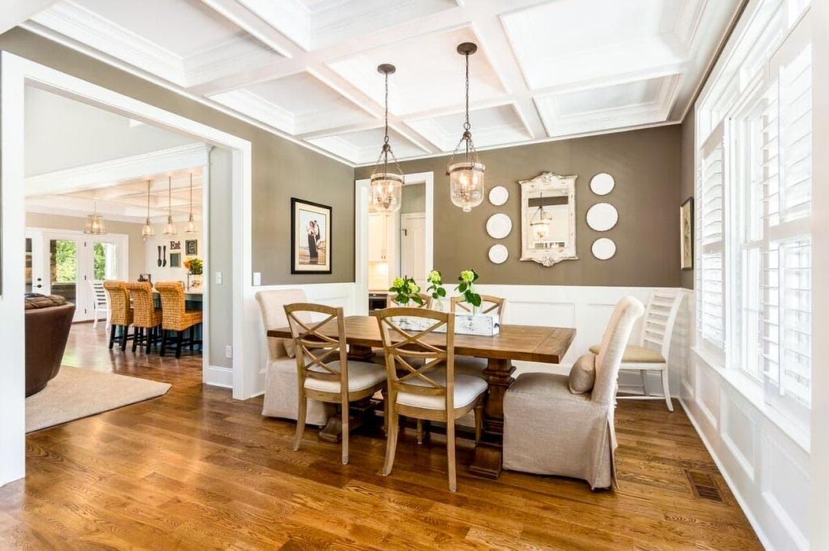 Dining room with coffered ceiling, wooden table and chairs, view into kitchen with island seating and bar stools.