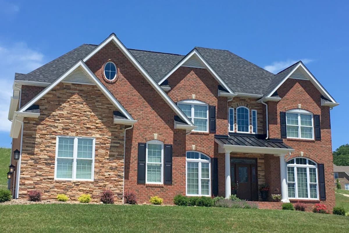 House plan exterior. Two-story brick home with gables, multiple dormers, and a front porch with columns. Features an oval window.