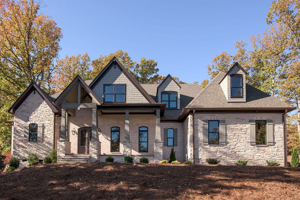 House plan exterior featuring a two-story design with a front porch, multiple gables, and dormers. Stone facade and shingle roofing are visible.
