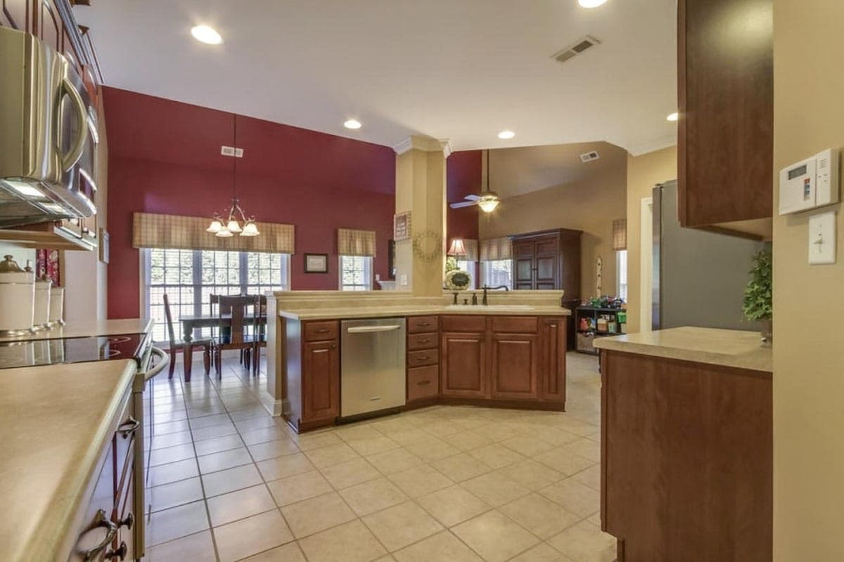 Kitchen with wooden cabinets, stainless steel dishwasher, tiled floor, and dining area with large windows.