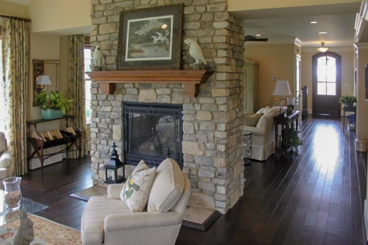 Interior view of a great room with a large stone fireplace, wooden mantel, and access to a hallway with a front door.