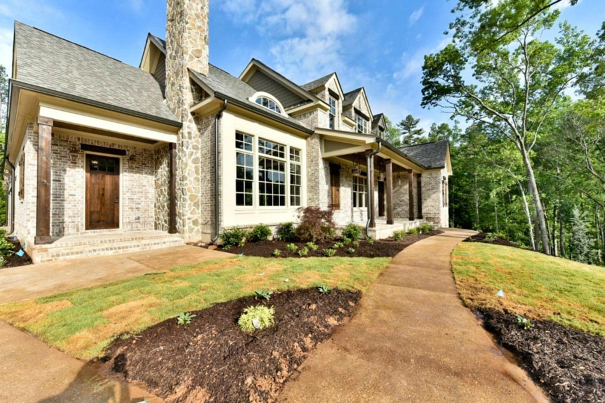 House plan exterior: Two-story home with brick facade, covered porch supported by timber posts, stone chimney, and multiple gables.