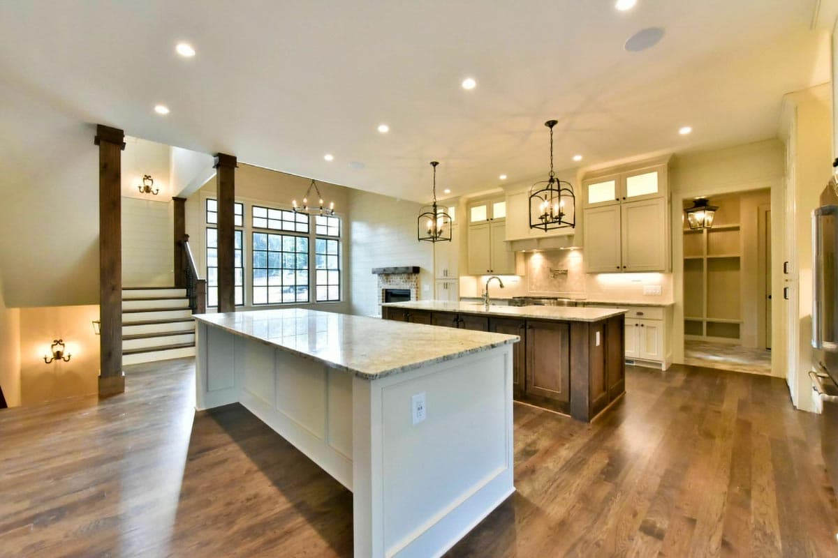 Open-concept kitchen with two islands, fireplace, large windows, and exposed wood beams on staircase.