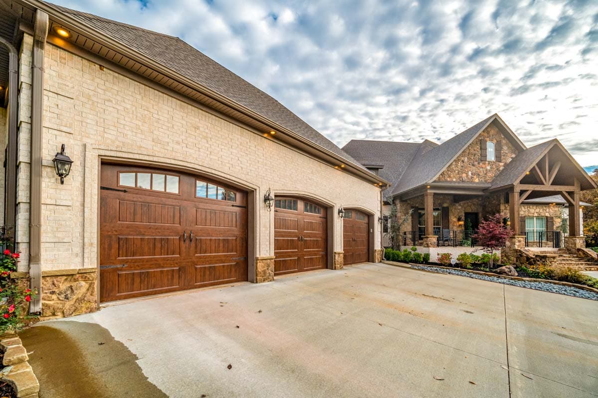 House plan exterior with light brick facade, three wooden garage doors, stone accents, and covered entry with timber framing.