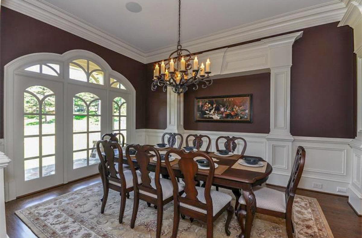 Dining room with arched French doors, ornate chandelier, wainscoting, and a large wooden table set for eight.