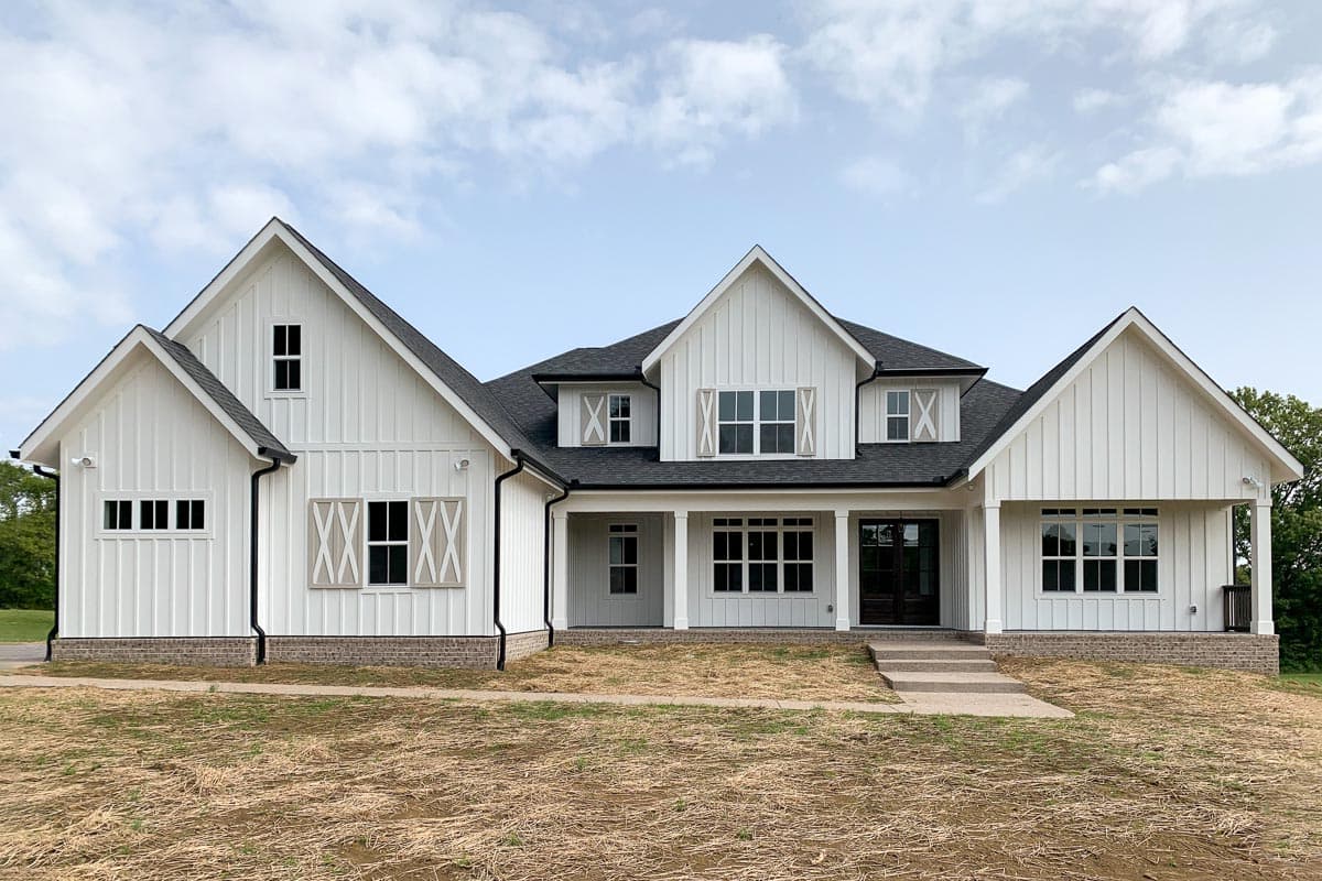 House plan exterior. Two-story Modern Farmhouse with gables, porch, and a dark roof. Features window shutters and a brick foundation.