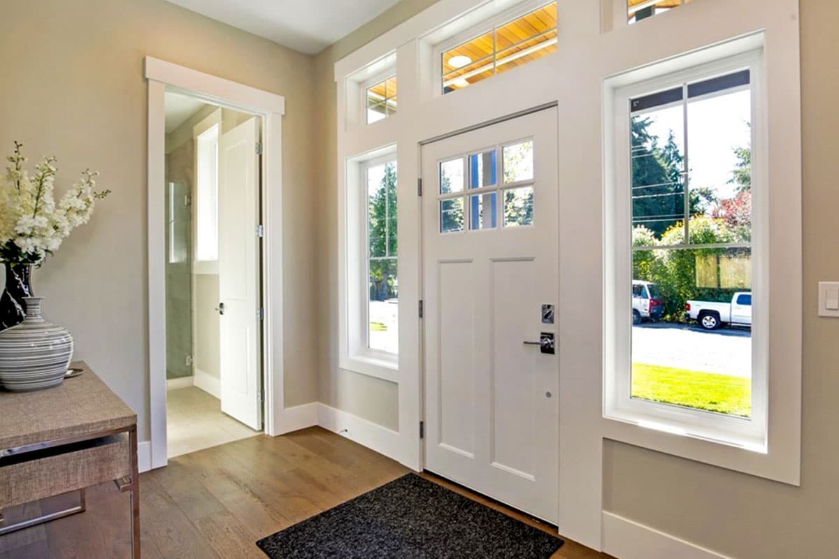 Interior view of a home entryway featuring a white paneled door with glass panes, flanking windows, and a door leading to a hallway.