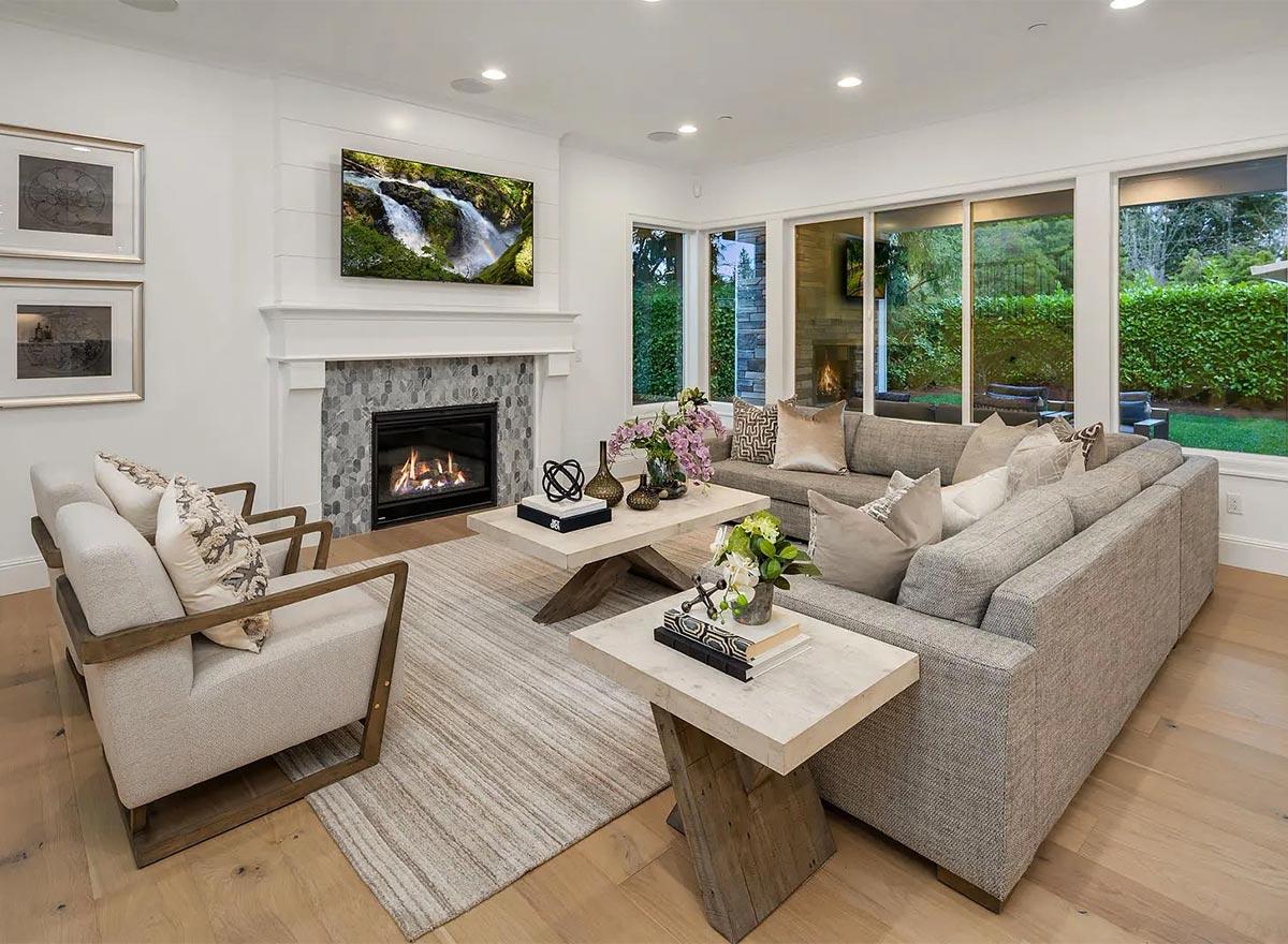 Living room interior with fireplace, TV, sectional sofa, and large windows overlooking a backyard.