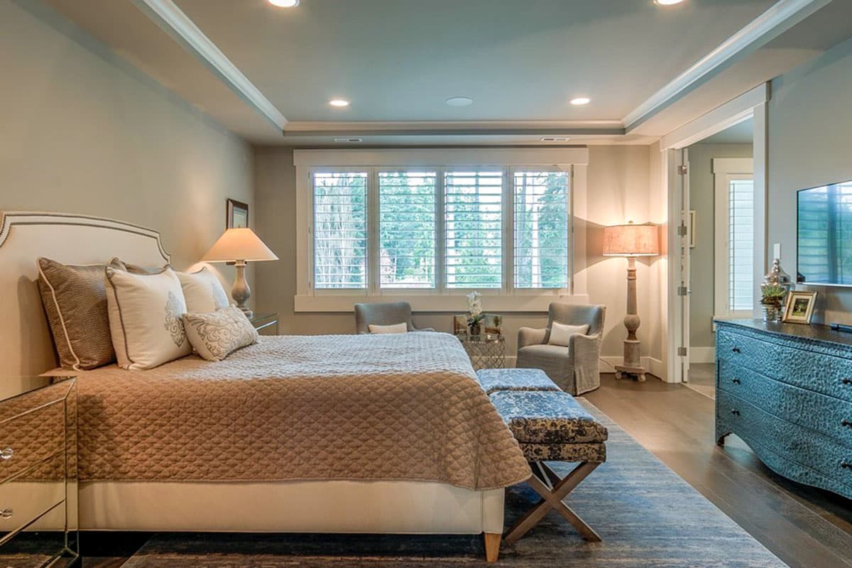Primary bedroom with tray ceiling, large windows with plantation shutters, tufted headboard, and accent seating.