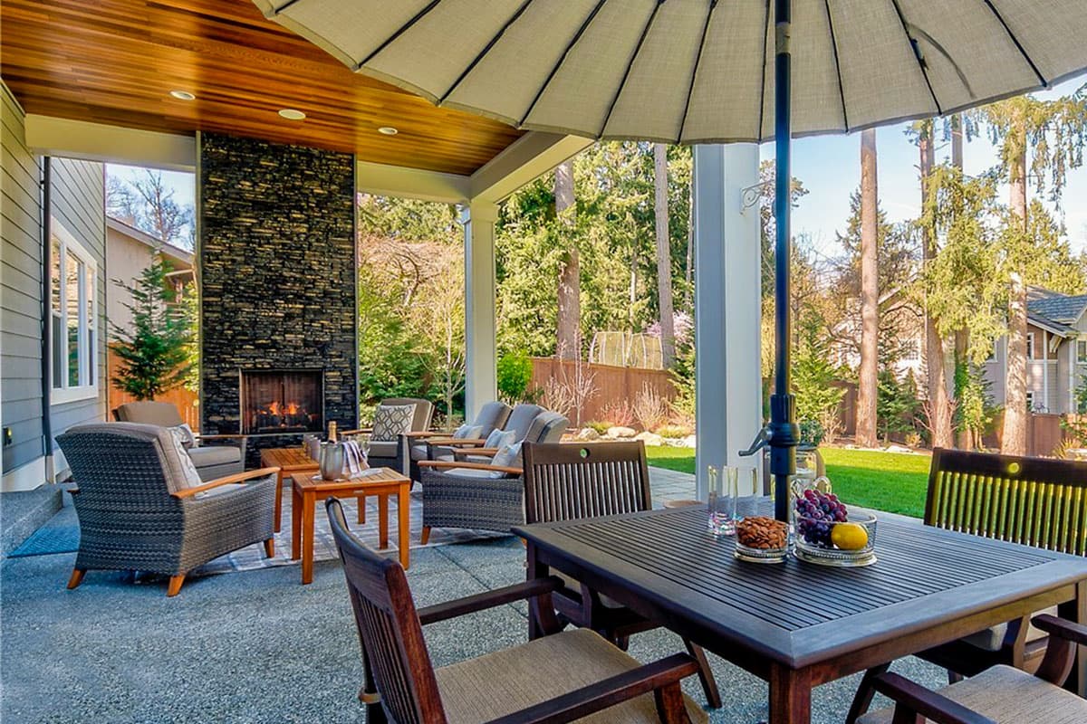 Covered patio with stone fireplace, seating area, and outdoor dining table with trees in background.