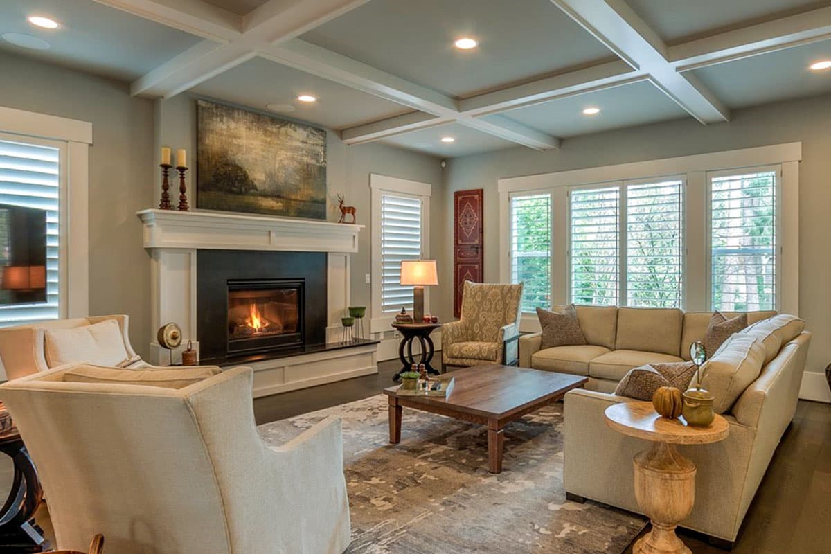 Living room with coffered ceiling, fireplace, seating area, and large windows with shutters.