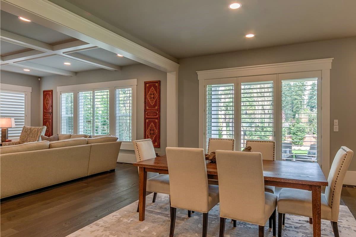 Interior view of a great room and dining area with coffered ceiling, large windows with shutters, and wooden flooring.