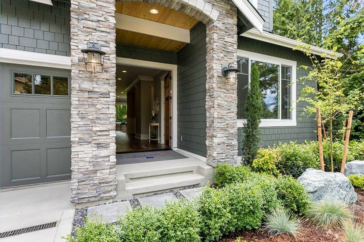 House plan exterior featuring a stone-clad arched entryway, gray garage door, and green shake siding.
