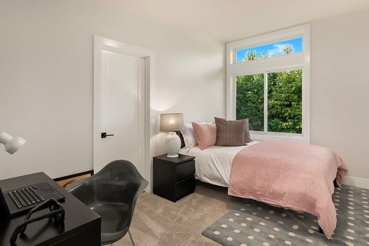 Bedroom interior with a bed, nightstand, desk, chair, and large window overlooking trees.