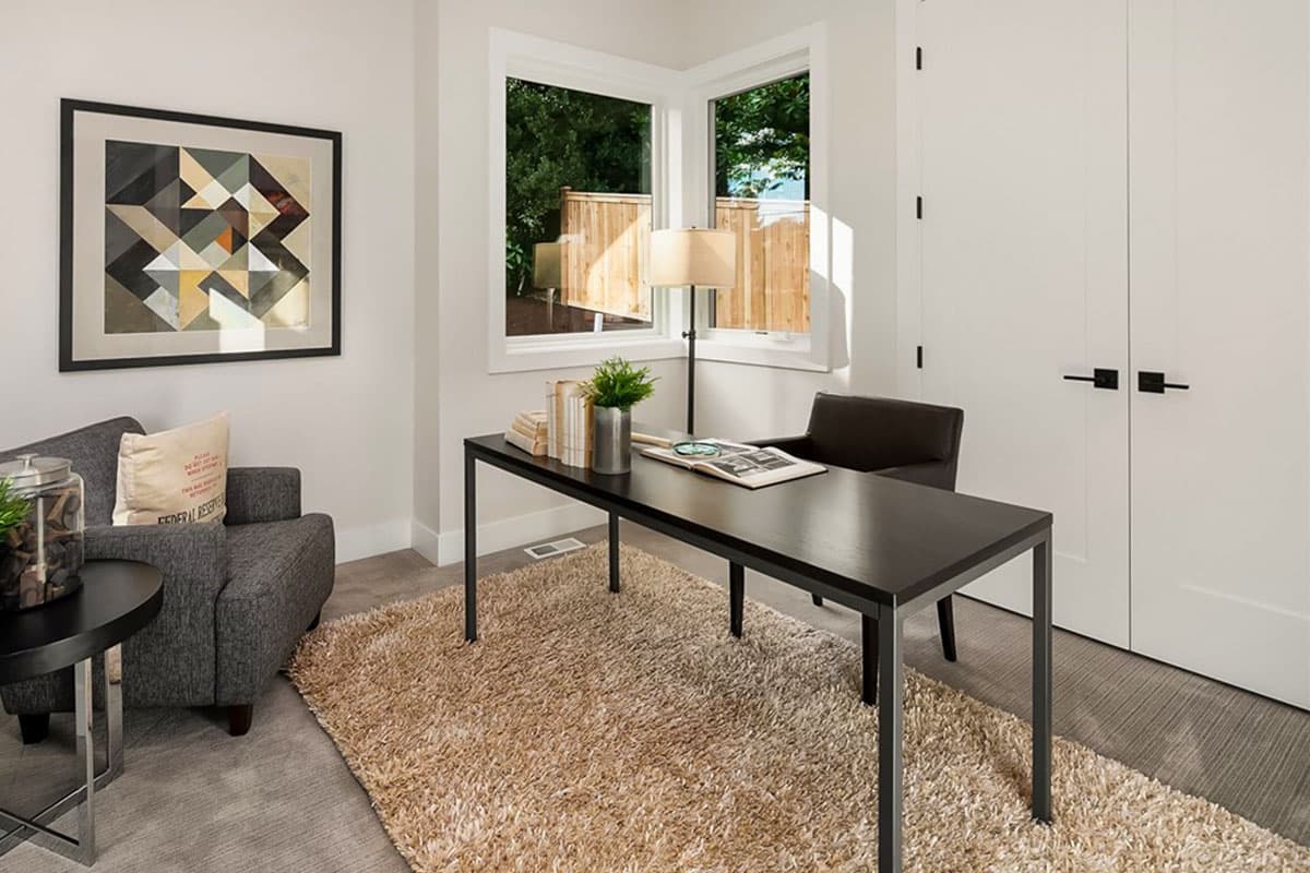 Home office with desk, chair, armchair, and corner windows overlooking a wooden fence.