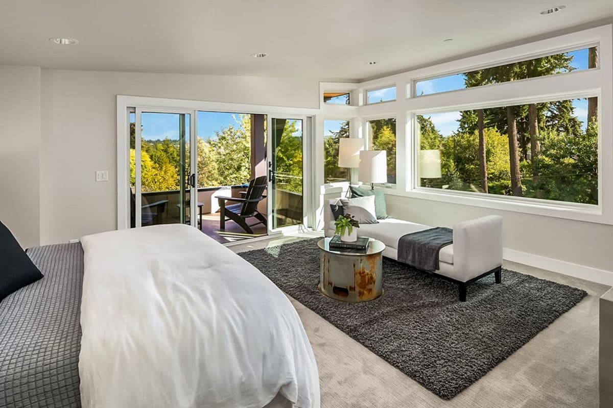 Bedroom with large windows, sliding glass door to balcony, and scenic wooded view.