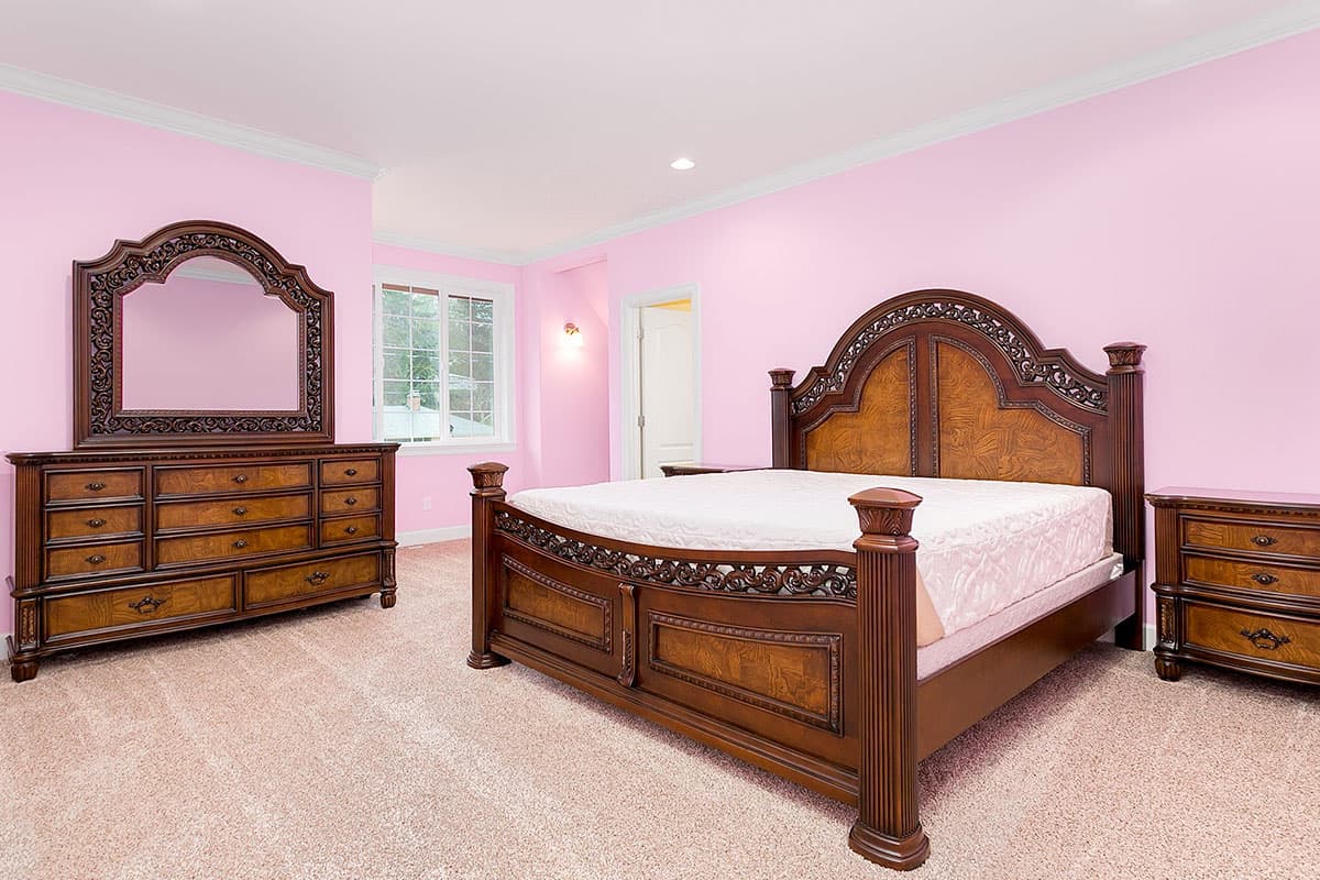 Bedroom with ornate dark wood furniture, including a king bed, dresser with mirror, and nightstands. Pink walls and carpet.