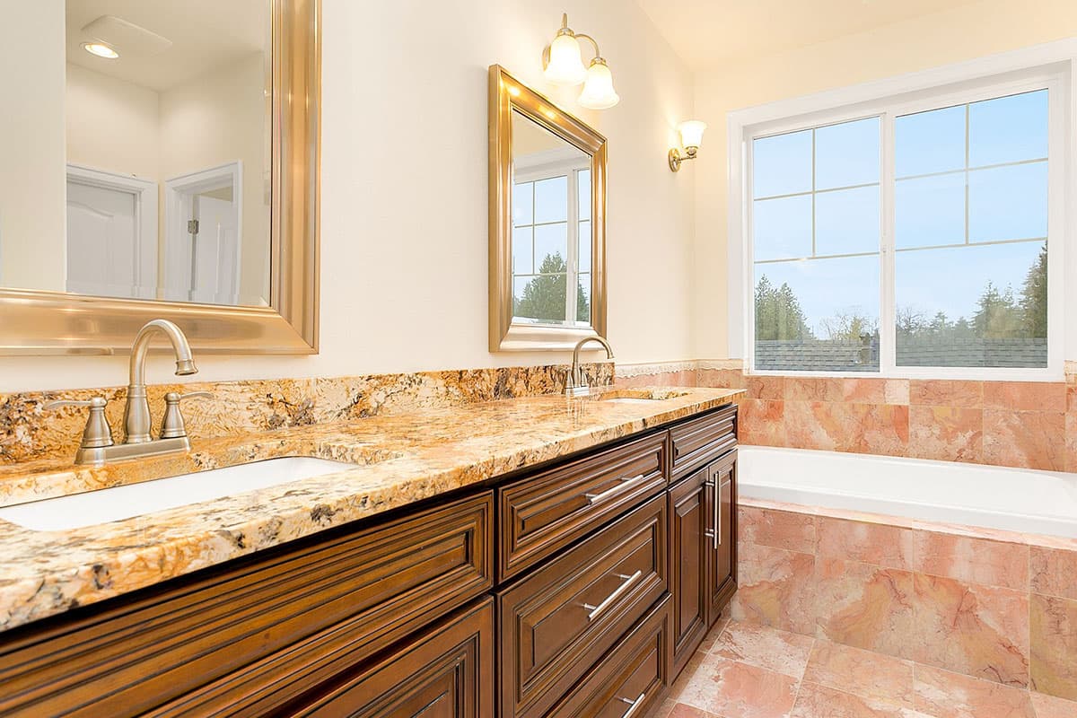 Master bathroom with double vanity, granite countertops, wood cabinets, large window, and tiled tub surround.