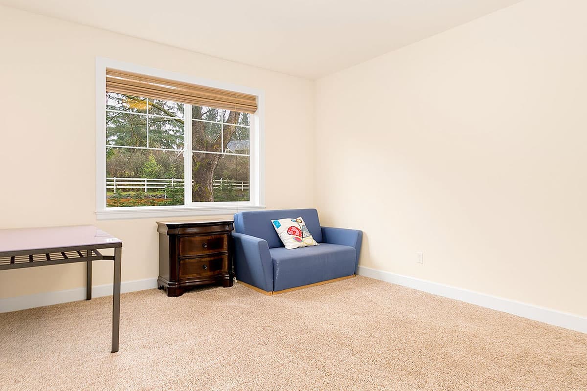 Interior view of a room with a blue loveseat, wooden nightstand, metal desk, and large window overlooking trees.