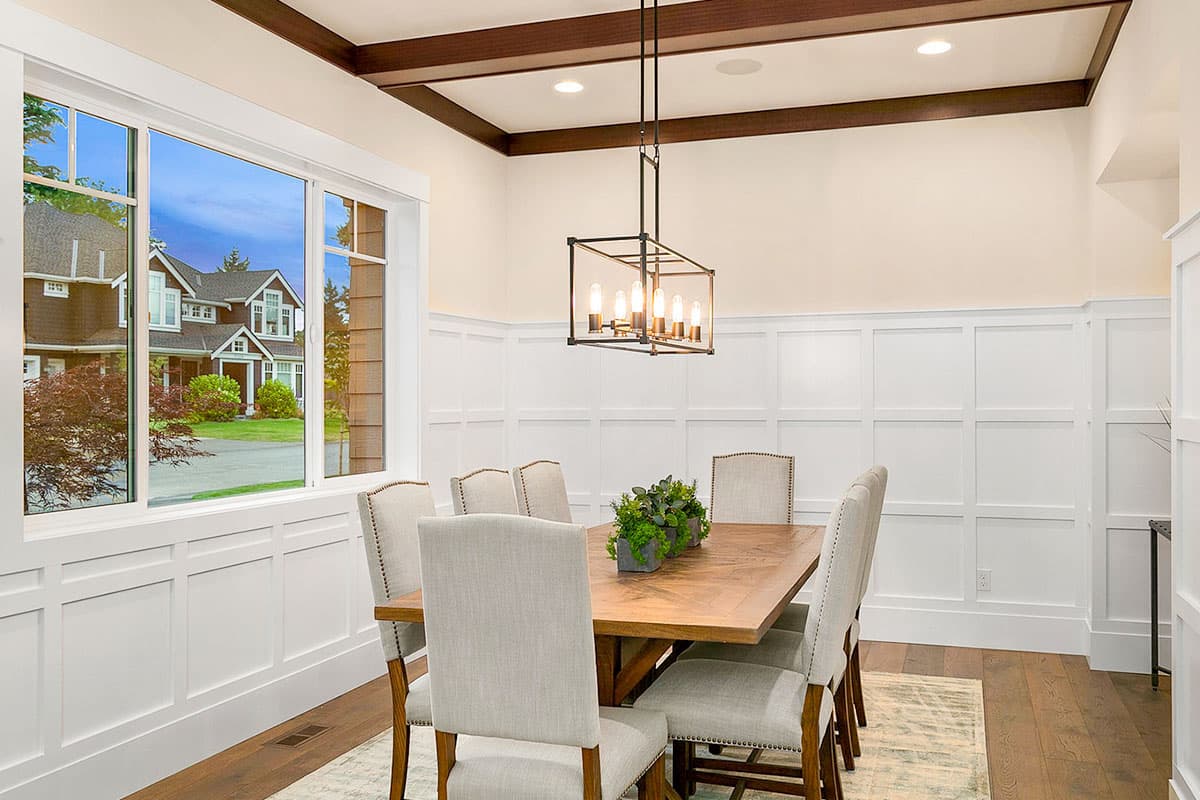 Dining room with wood ceiling beams, wainscoting, and a large window overlooking a Craftsman-style house.