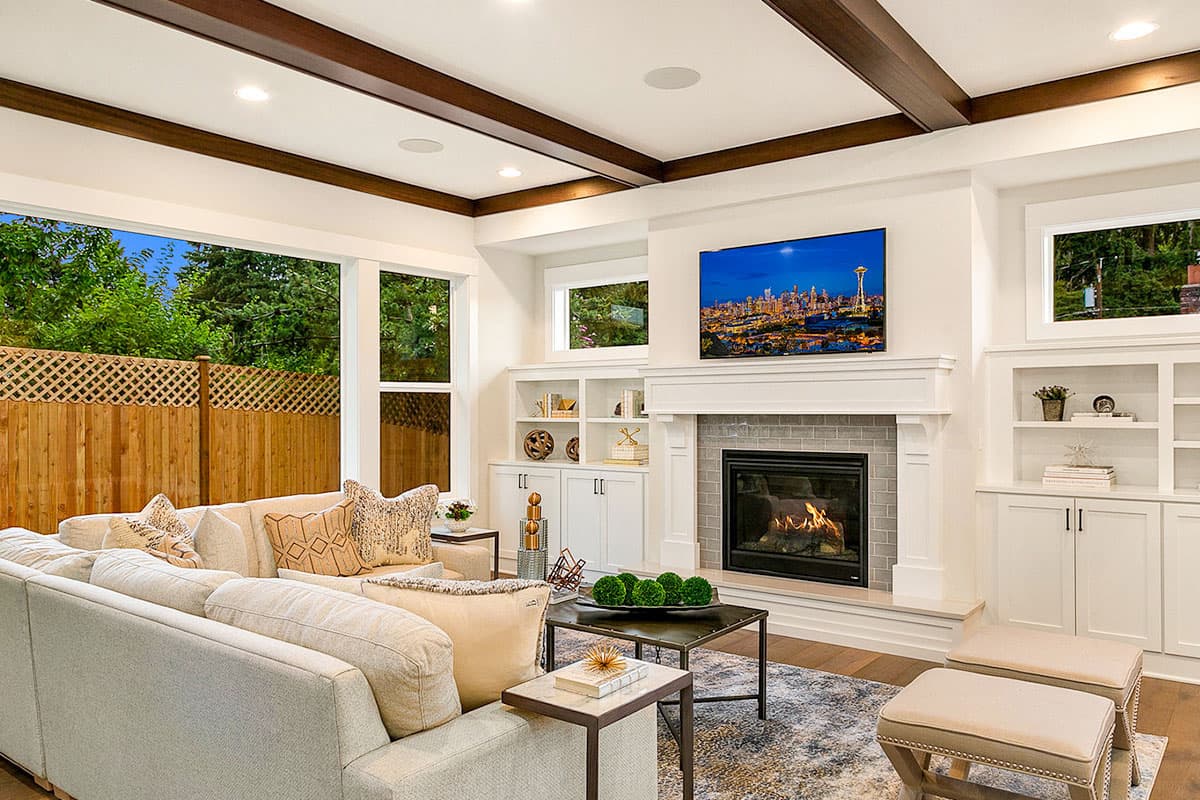 Great room with fireplace, built-in shelving, coffered ceiling, and large windows overlooking a backyard.
