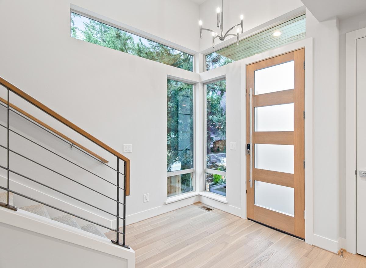 Modern entryway with open staircase, wood-paneled door, and floor-to-ceiling corner windows.