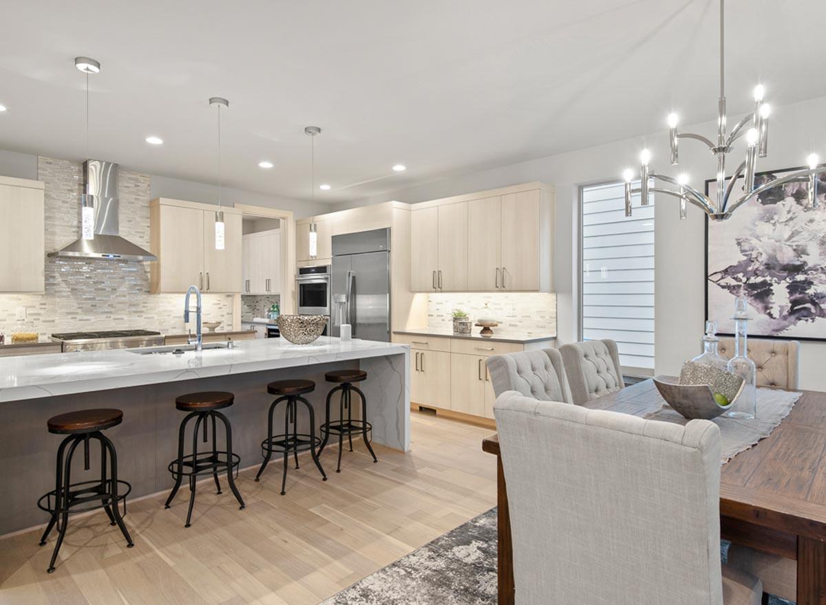 Modern kitchen with light wood cabinetry, large island with seating, stainless steel appliances, and adjacent dining area with upholstered chairs.