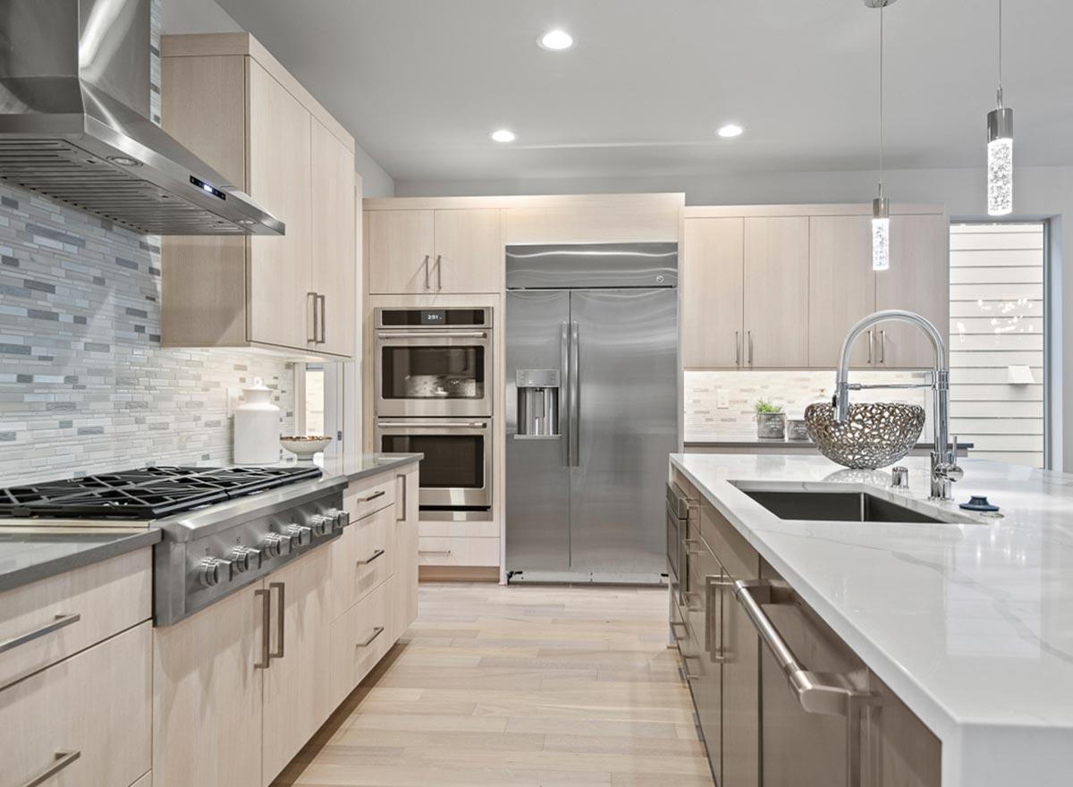 Modern kitchen with light wood cabinets, stainless steel appliances including a double oven and refrigerator, and a large island with a sink.