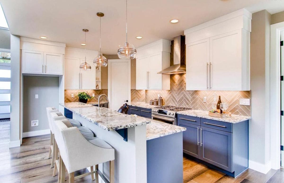 Modern kitchen with white upper cabinets, blue lower cabinets, granite island, stainless steel range hood, and herringbone backsplash.