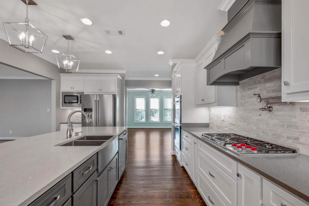 Kitchen with large island, stainless steel appliances, white cabinets, gray vent hood, and tile backsplash.