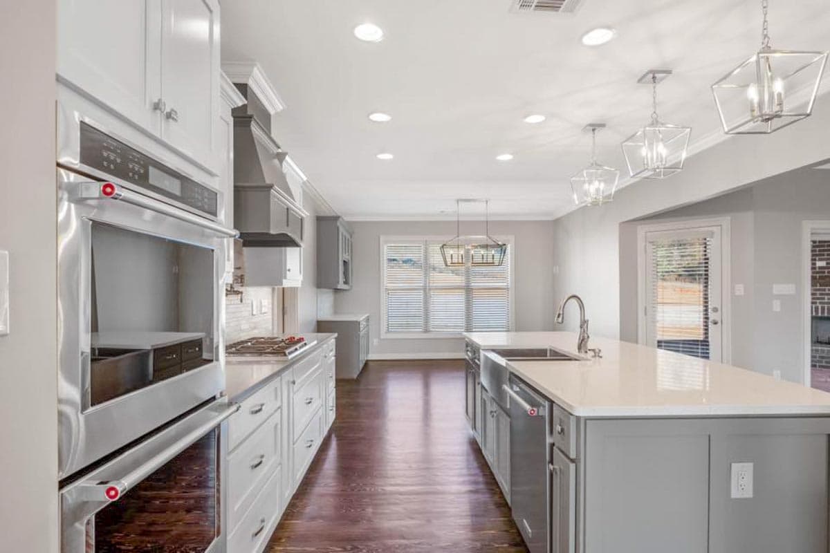 Kitchen with double ovens, gray island with sink, white counters, pendant lights, and hardwood floors.