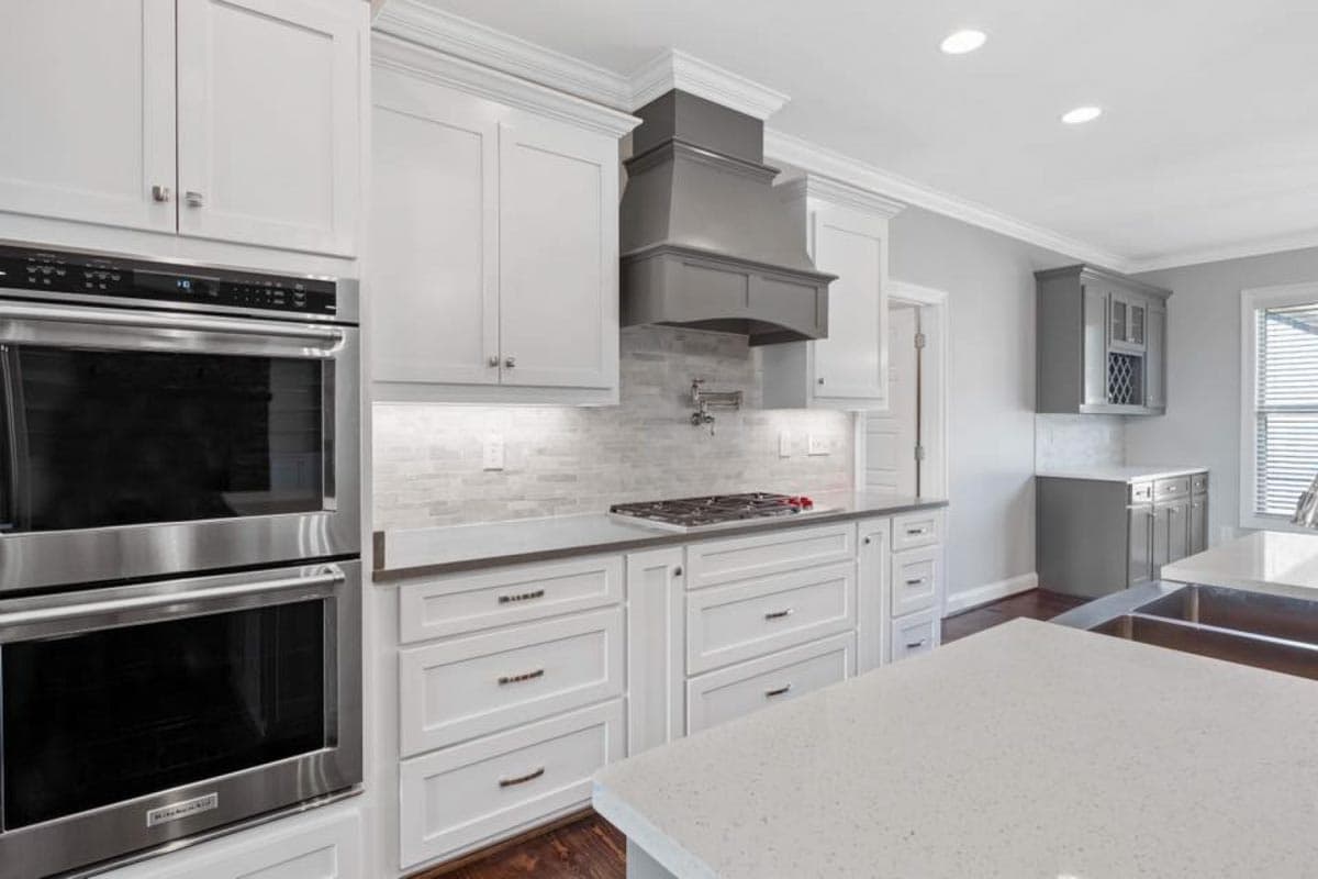 Kitchen with white Shaker cabinets, dual ovens, gas range, gray vent hood, marble tile backsplash, and quartz countertops.