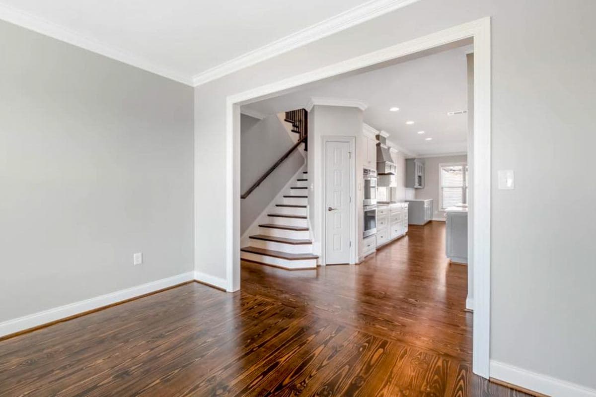 Interior view of a house showing a staircase leading up, a kitchen with white cabinets and appliances, and dark hardwood floors.