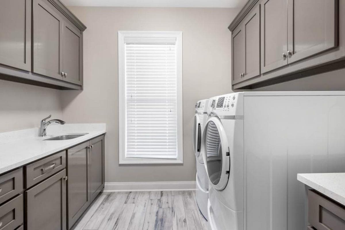 Laundry room interior with gray cabinetry, white quartz countertop, sink, washer, dryer, and window with blinds.