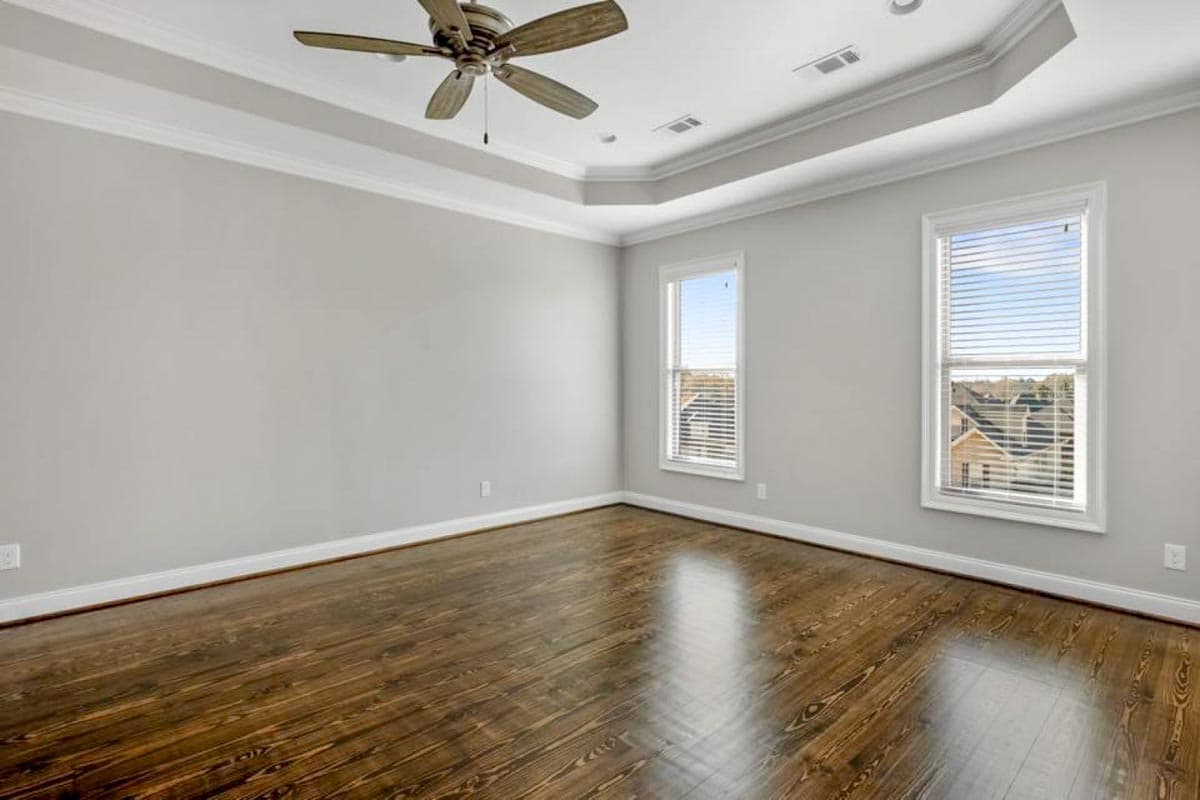 Interior view of a bedroom with a tray ceiling, ceiling fan, and two windows with blinds. Features dark wood floors and light gray walls.