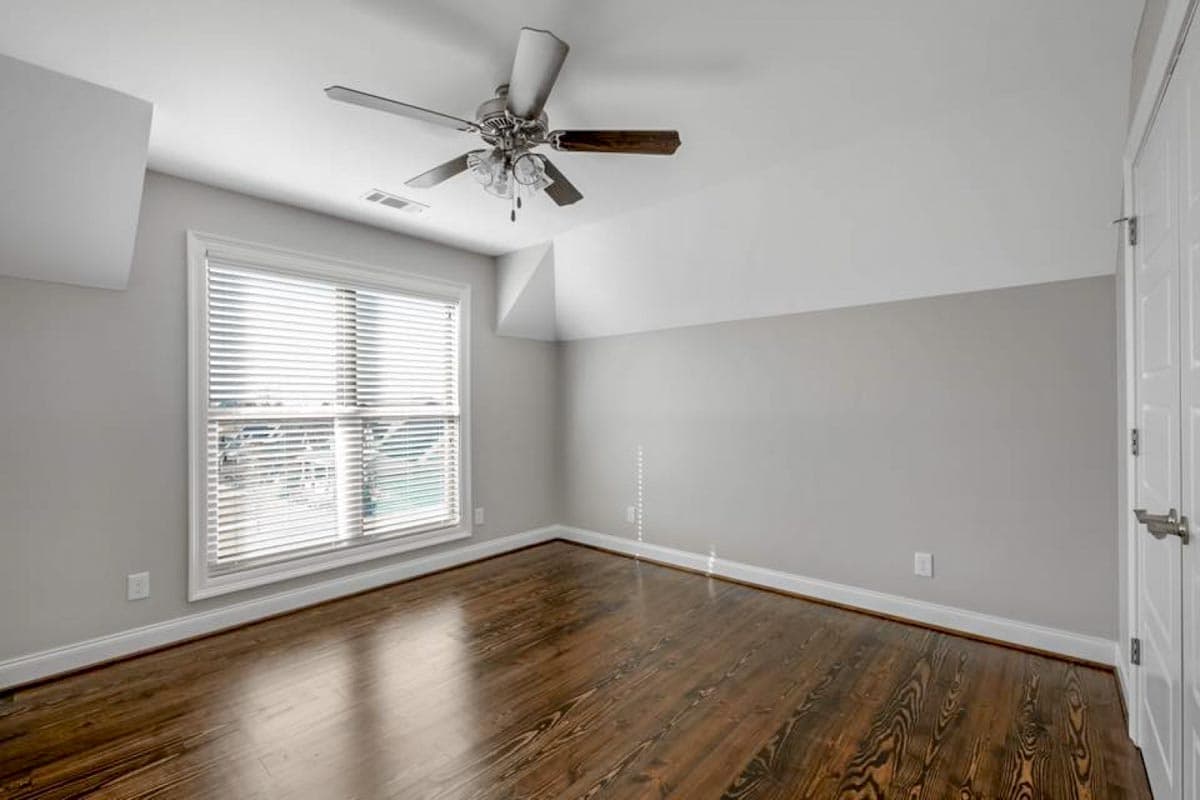 Interior view of an empty room with dark hardwood floors, a ceiling fan, and a large window with blinds.