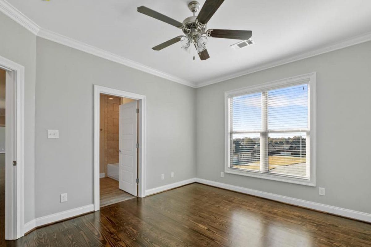 Bedroom with hardwood floors, ceiling fan, and doorway to bathroom. Large window with view of neighborhood.