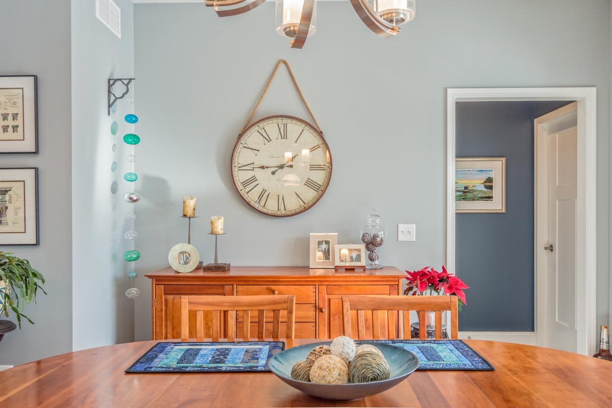 Dining room featuring a wooden buffet with a large wall clock, two chairs with placemats on a table.