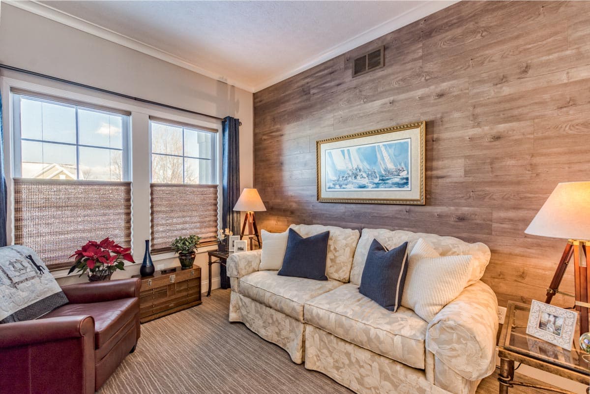 Living room with wood-paneled accent wall, floral sofa, leather armchair, and two windows with blinds.