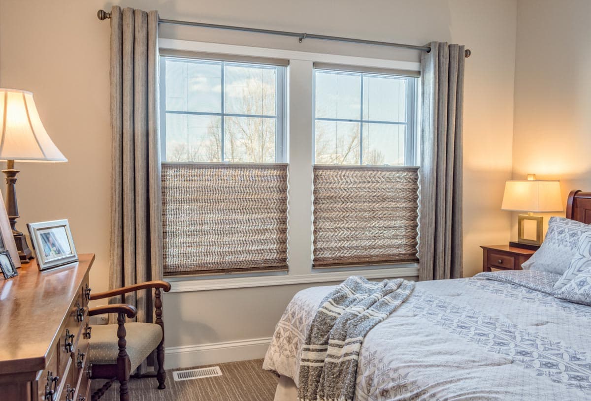 Bedroom interior with two windows featuring textured blinds, draped curtains, dresser, chair, and bed with patterned bedding.
