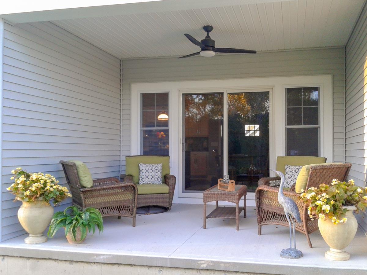 Covered porch with siding, ceiling fan, sliding glass doors, and two chairs with cushions.
