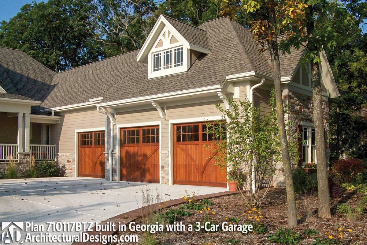 House plan exterior: Beige siding, stone accents, gable dormer with multiple windows, and three wooden garage doors.